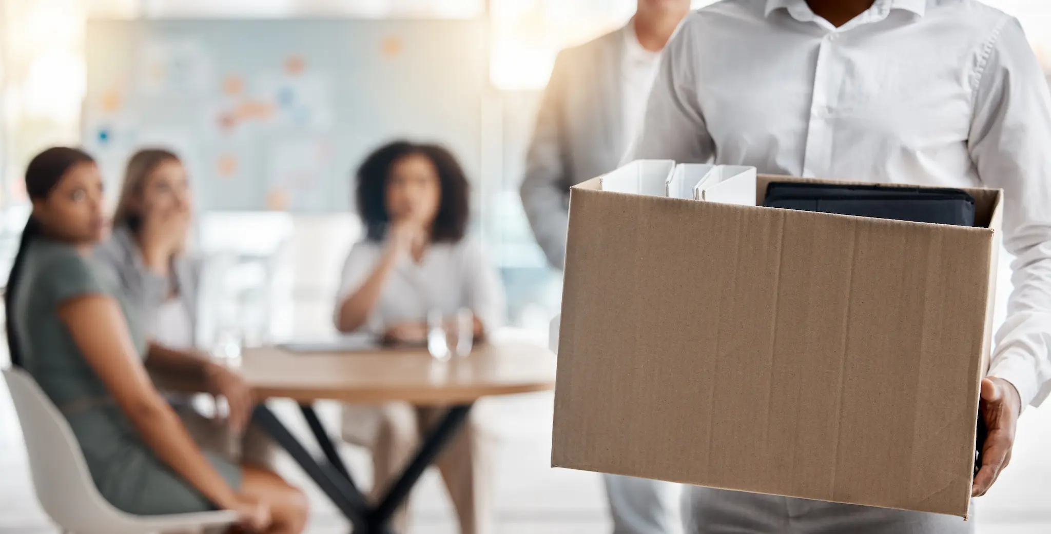 Persona llevando una caja con objetos personales tras un despido disciplinario, con colegas al fondo observando.