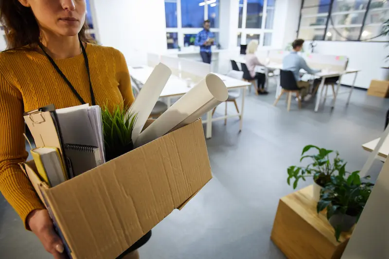 Mujer llevando una caja con pertenencias personales tras un despido en una oficina moderna.