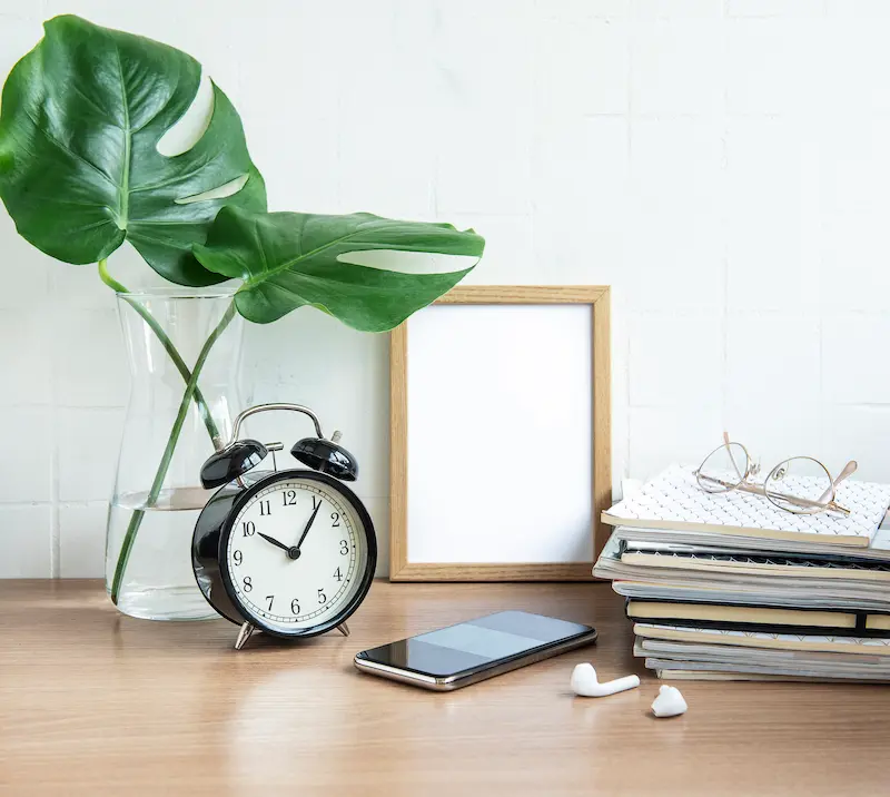 Reloj despertador negro junto a hojas verdes en jarrón de vidrio, teléfono móvil y libros apilados sobre escritorio.